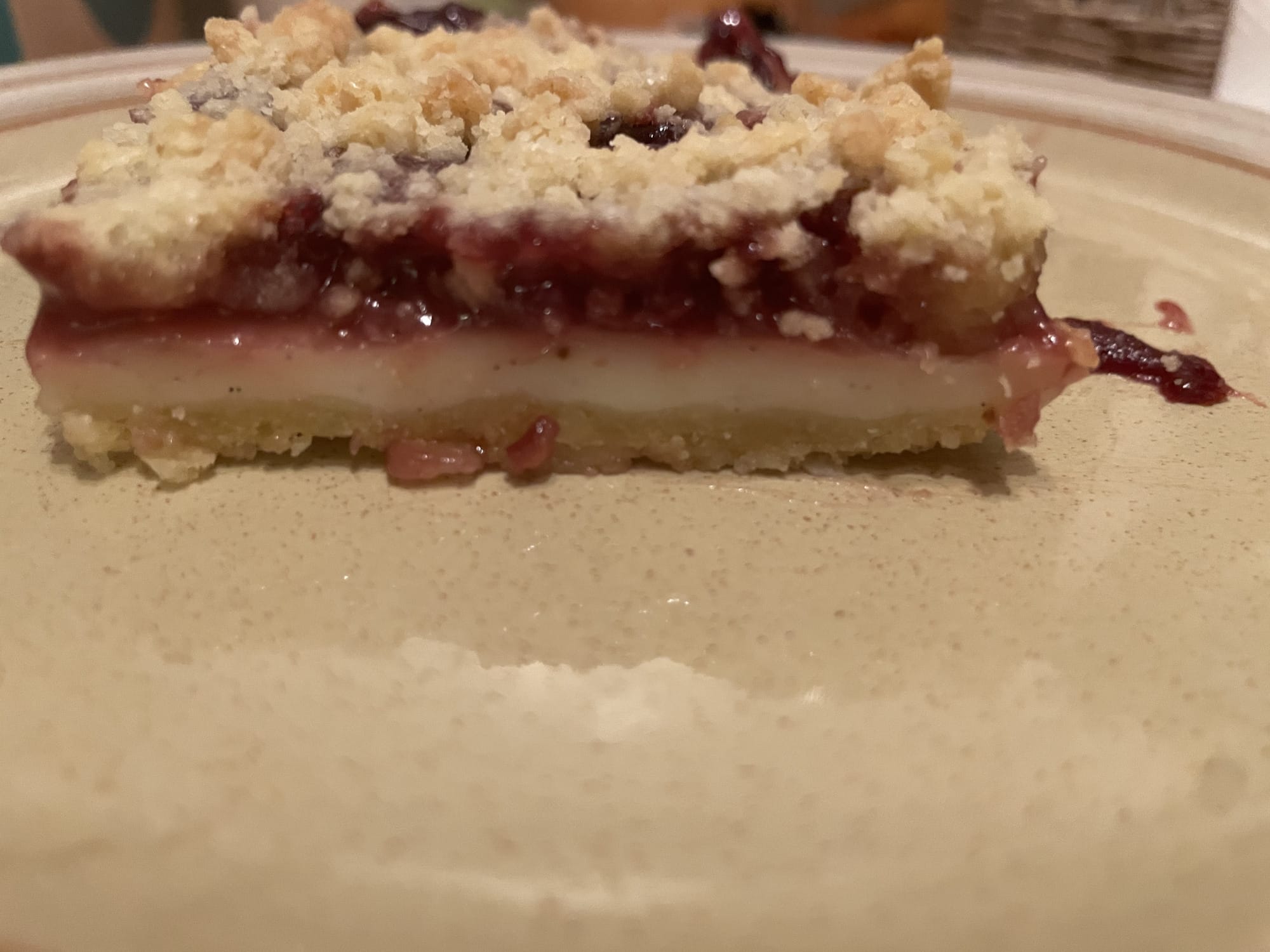 Ein Bild des Kuchens, bei dem man besonders gut die verschiedenen Lagen sieht. Ganz oben crumbles, darunter Kirschen, dann die weiße Vanillesoße und ganz unten der crumbles Boden. Das Kuchenstück liegt auf einem Teller.