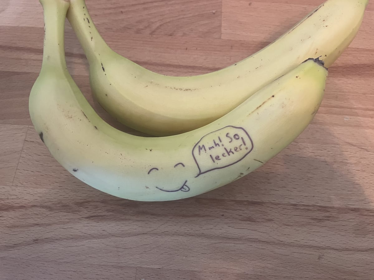 2 gelbe Bananen. Auf der vordersten ist ein Gesicht mit geschlossenen Augen und einem lächelndem Mund der sich um den Mund leckt. Eine Sprechblase ist daneben die sagt: Mmh! So lecker!