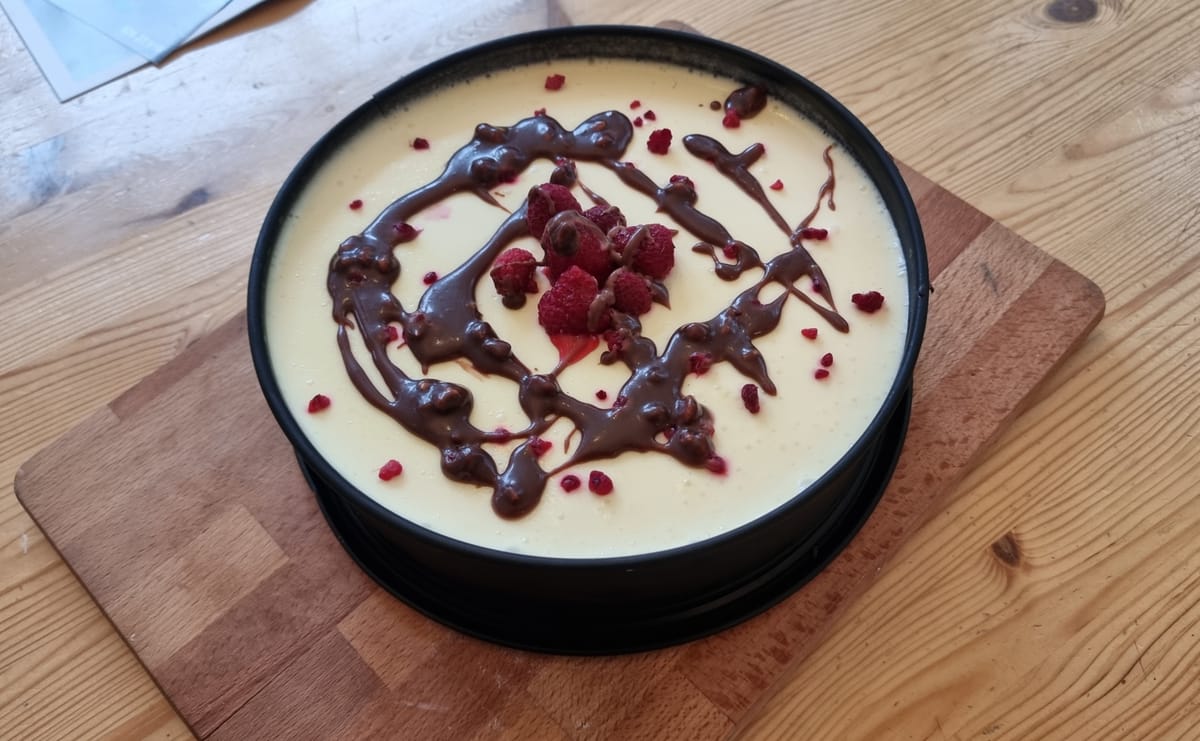 Ein Bild von einem Käsekuchen in einer Kuchenform. Der Kuchen steht auf einem Schneidebrett auf einem Tisch. Er ist mit Himbeeren und geschmolzener Schokolade faziert. 
